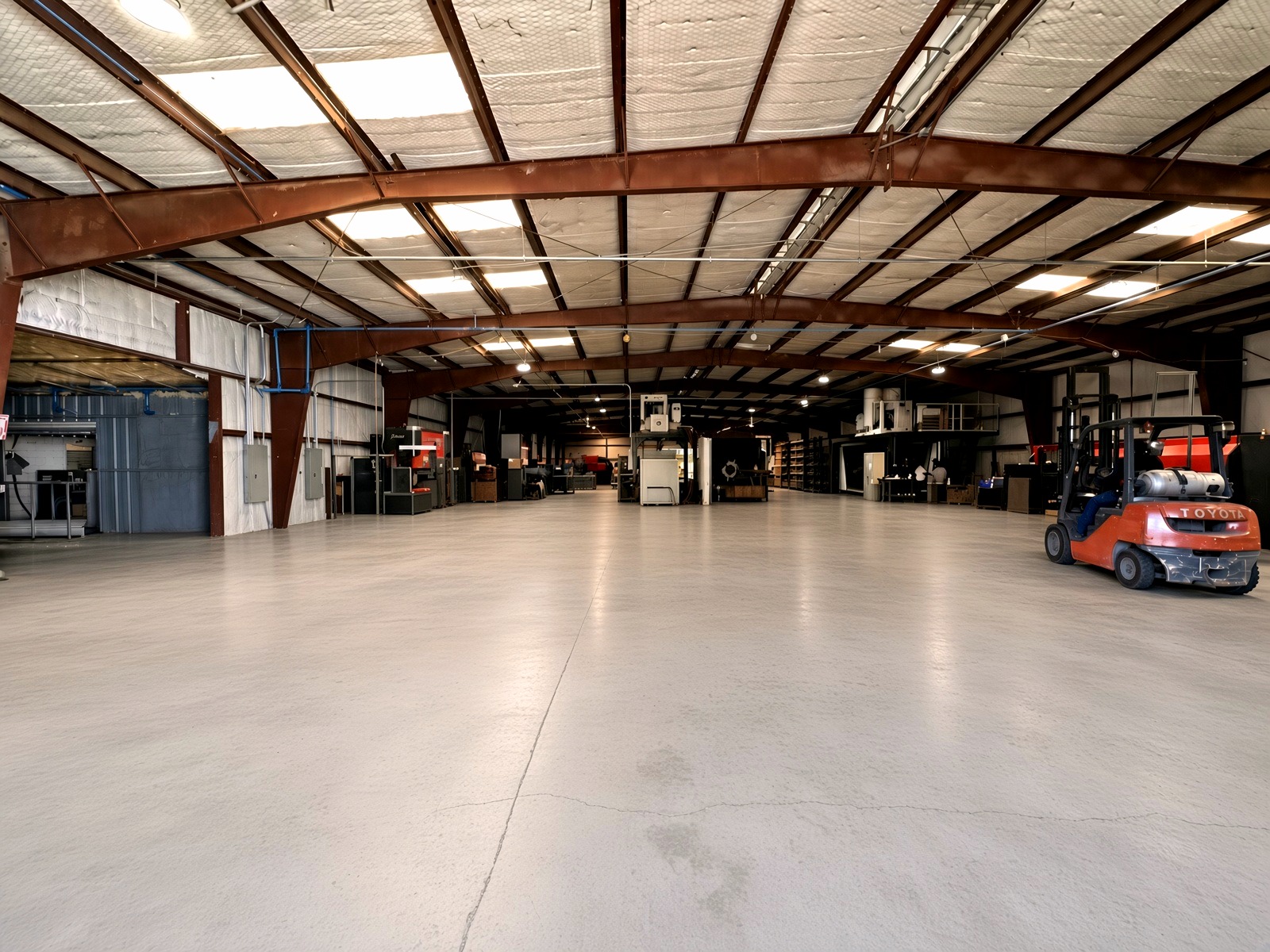 Wide interior view of the Top J Metal Works production floor with red steel beams, overhead lighting, and a Toyota forklift